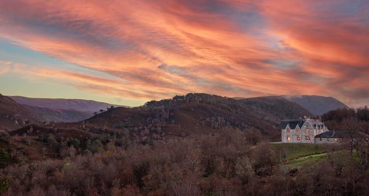 Casa señorial en una ladera boscosa bajo unas nubes de atardecer dramáticas de color rosa y naranja.