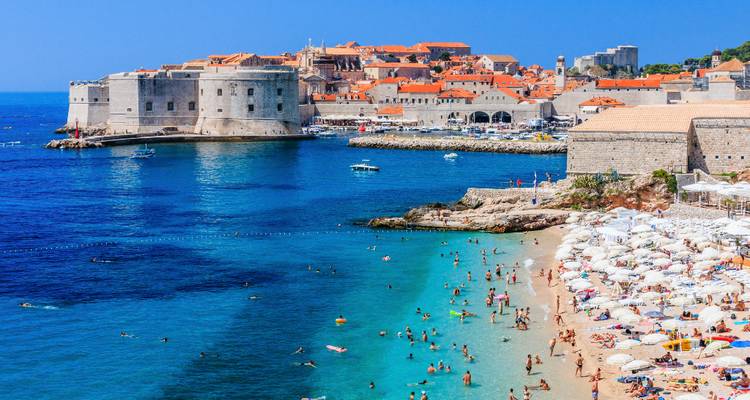 Vue aérienne vibrante de la vieille ville fortifiée de Dubrovnik et d'une plage turquoise animée en dessous.