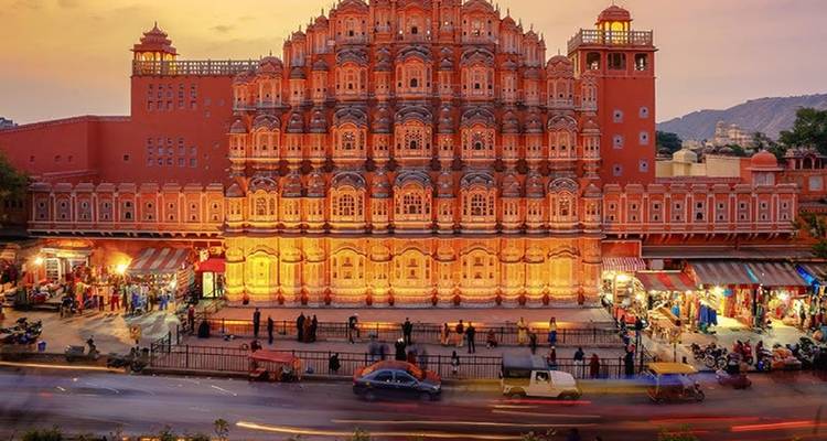 Beleuchtete Fassade des Hawa Mahal von Jaipur, die bei Sonnenuntergang leuchtet, mit lebhaftem Straßenverkehr darunter.
