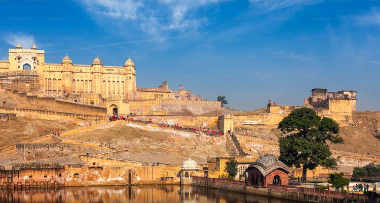 Panoramablick auf die hellgelben Wälle der Amber Fort, die sich über dem See und der Hanglage unter blauem Himmel erheben.