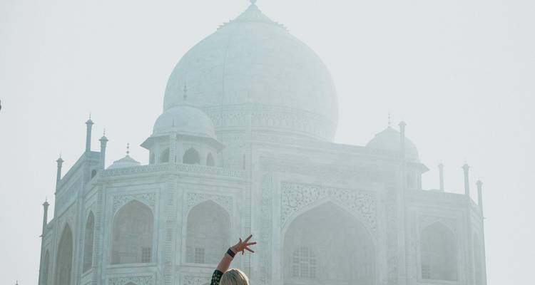 Ein Reisender, der vor dem Taj Mahal steht, teilweise in Morgennebel gehüllt, und freudig eine Hand hebt.