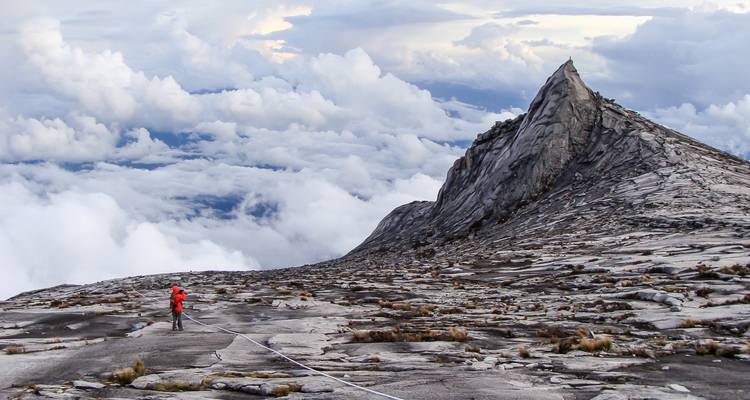 Einzelner Wanderer in einer roten Jacke überquert das Granitgipfelplateau des Mount Kinabalu über den Wolken.