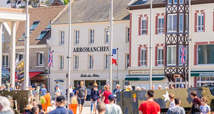Scène de rue à Arromanches avec drapeaux, cafés et touristes commémorant l'histoire du Débarquement sous un ensoleillement radieux.
