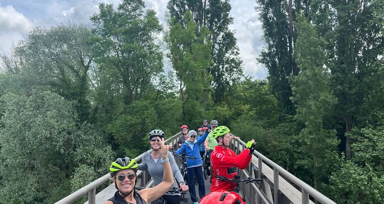Des cyclistes souriants portant des casques posent sur un étroit pont en bois entouré d'une végétation luxuriante.