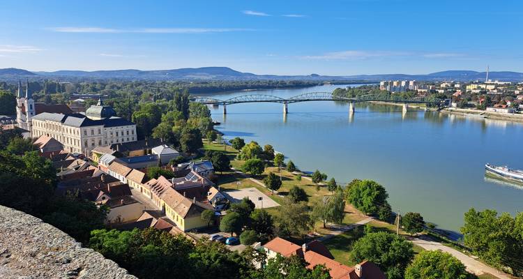 Vista panorámica de un pueblo ribereño con una basílica y un puente de arcos que cruza el Danubio en un día despejado.