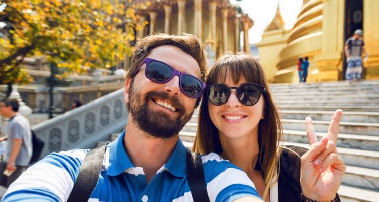 Un couple souriant prend un selfie devant les marches ornées d'un temple doré, faisant le signe de la paix.