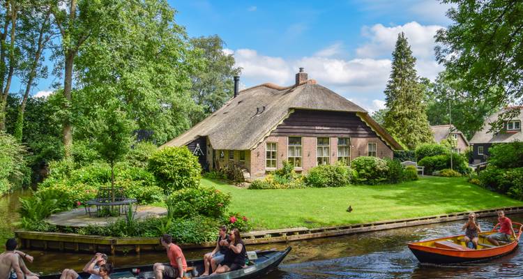 Reetgedecktes Cottage mit üppigem Garten an einem Kanal in Giethoorn, während kleine Boote mit Besuchern vorbeigeleitet werden.