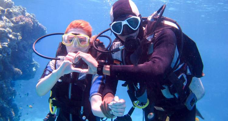 Dos buceadores haciendo una forma de corazón con sus manos junto a un arrecife de coral.