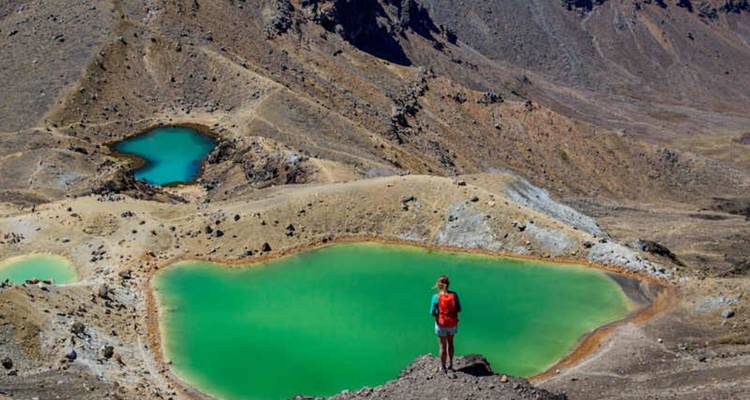 Un randonneur solitaire portant un sac à dos rouge se tient au-dessus des spectaculaires lacs d'Émeraude verts dans un paysage alpin volcanique.