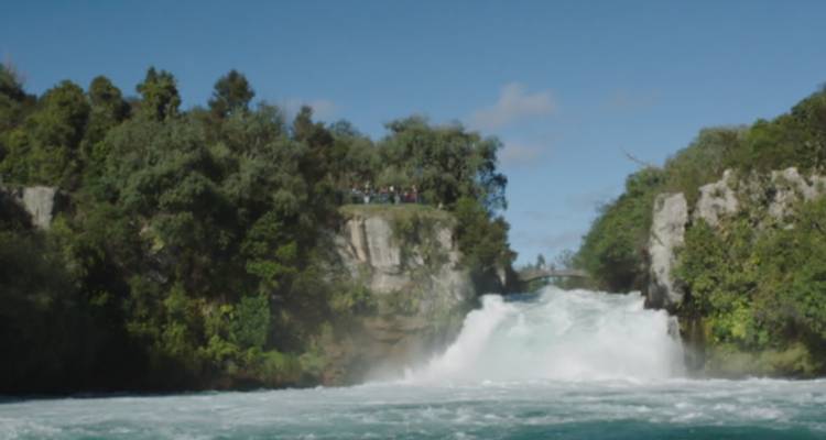 L'eau turquoise puissante se précipite sur les chutes de Huka tandis que des visiteurs lointains observent depuis un pont au-dessus.