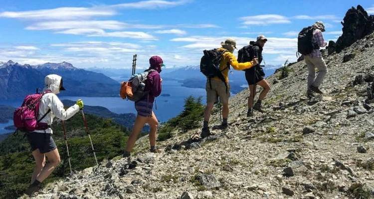 Cinq randonneurs gravissent une crête montagneuse rocheuse surplombant un lac bleu profond et des pics patagoniens enneigés sous un ciel dégagé.