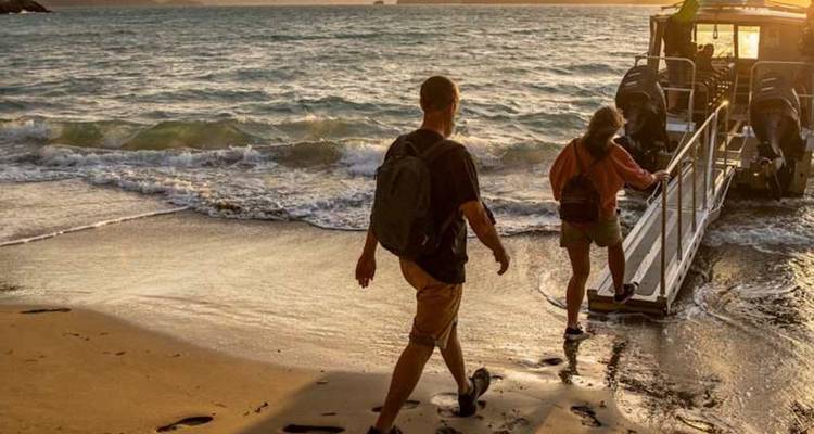 Les voyageurs montent à bord d'un petit bateau depuis une plage de sable fin au coucher de soleil doré, avec de légères vagues qui clapotent doucement.
