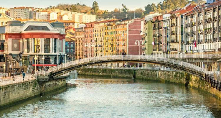 Puente peatonal elegante que cruza el río con los coloridos edificios históricos de Bilbao a lo largo de las orillas