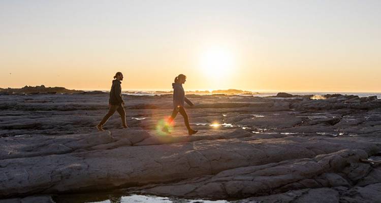 Deux voyageurs marchent sur les vasières rocheuses au lever du soleil, avec le soleil bas à l'horizon.