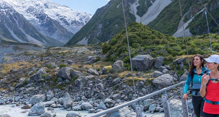 Les randonneurs font une pause sur un pont suspendu dans une vallée glaciaire encadrée par des pics enneigés.