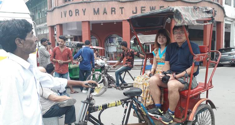 Touristen fahren mit einer Fahrradrikscha durch eine belebte Marktstrasse, die von Geschaeften und Fussgaengern gesaeumt ist.