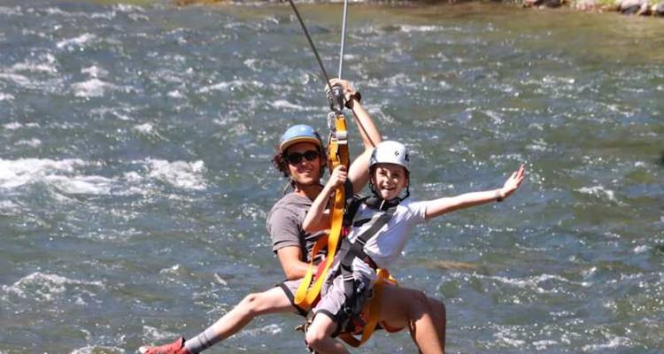 Un adulte et un enfant font de la tyrolienne ensemble, haut au-dessus d'une rivière qui coule.