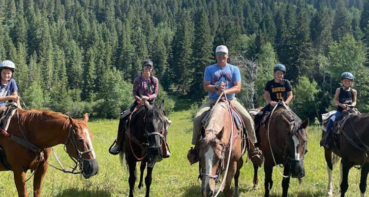 Une famille à cheval s'aligne dans une prairie ensoleillée bordée par une dense forêt de pins.