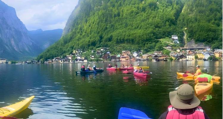 Grupos de kayakistas reman a través de un tranquilo lago alpino con el pintoresco pueblo de Hallstatt y escarpadas montañas verdes de fondo