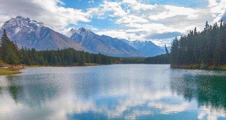 Lago alpino tranquilo que refleja picos cubiertos de nieve y bosques de coníferas bajo un cielo parcialmente nublado