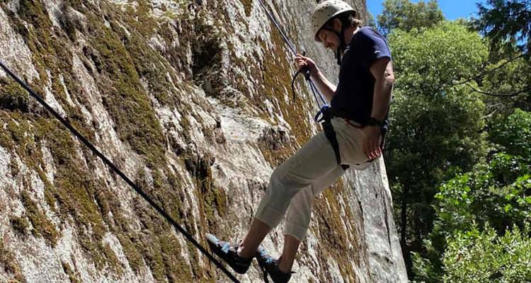 Un grimpeur portant un casque descend en rappel une paroi de granit couverte de mousse dans un canyon forestier.