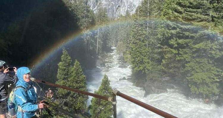 Un arc-en-ciel vibrant se dessine au-dessus d'une rivière tumultueuse et d'une forêt de pins tandis que des randonneurs emmitouflés observent la scène depuis un pont.