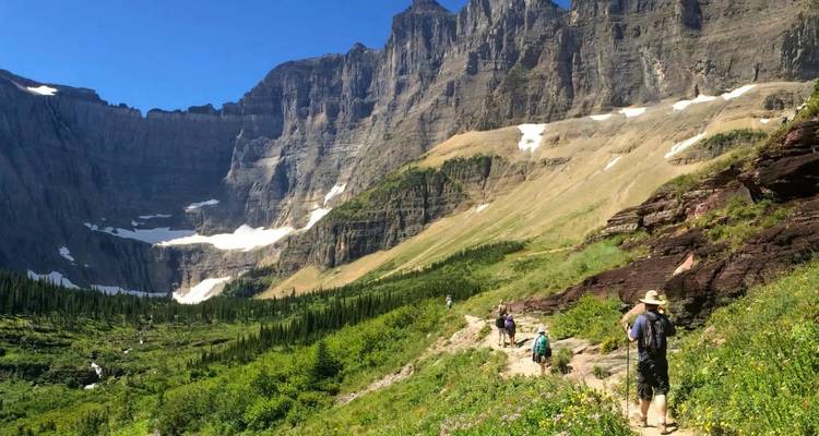 Los excursionistas recorren un exuberante sendero alpino bajo imponentes paredes de roca con parches de nieve que persisten.