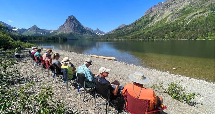 Un grupo grande se relaja en sillas de campamento a lo largo de la orilla tranquila de un lago de montaña.