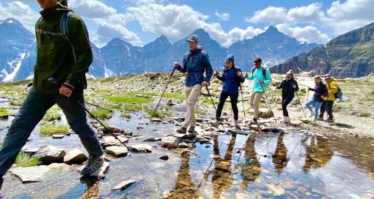 Los excursionistas con bastones cruzan un arroyo alpino poco profundo bajo picos cubiertos de nieve.