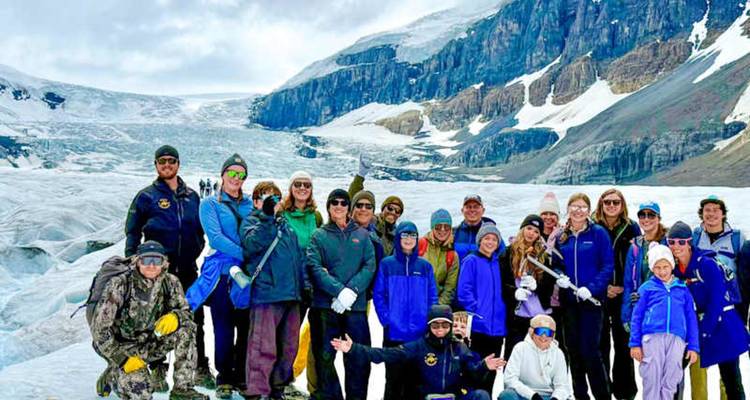 Un gran grupo de turistas sonríe en la cima de un glaciar extenso enmarcado por acantilados rocosos de las Montañas Rocosas.