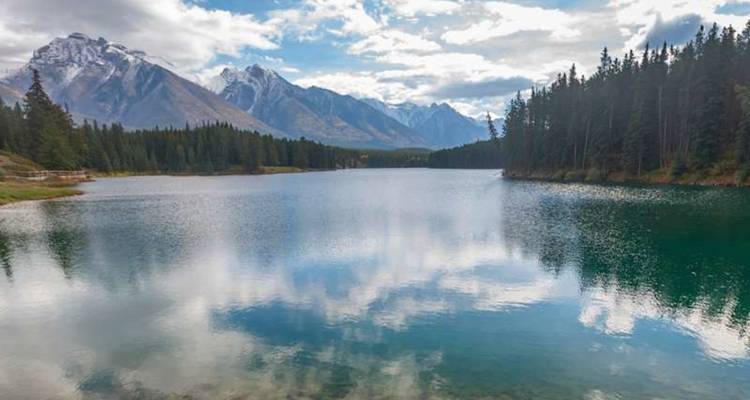 Un tranquilo lago alpino refleja picos nevados y nubes entre densos bosques de pinos.