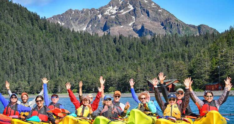 Un groupe de kayakistes joyeux lève les bras avant des montagnes côtières aux sommets enneigés.