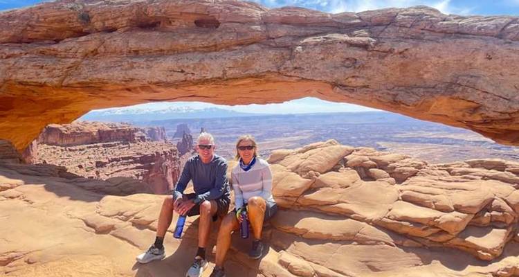 Deux voyageurs sont assis sous une arche naturelle de grès surplombant un vaste paysage de canyon sous un ciel bleu.
