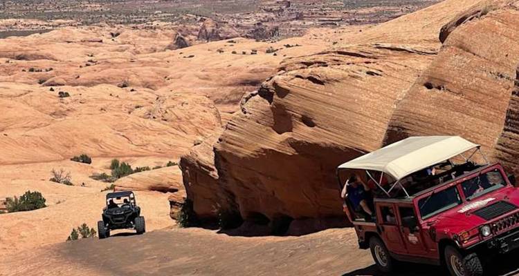 Des véhicules tous-terrains rouges et noirs gravissent une pente de désert de roche lisse parmi des formations de grès spectaculaires