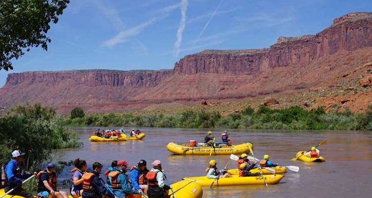 Plusieurs radeaux jaunes remplis de pagayeurs flottent sur une large rivière dominée par des parois de canyon rouge et un ciel bleu.