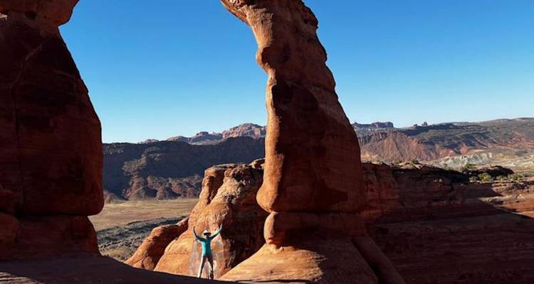 Une personne lève les bras sous l'imposante Delicate Arch encadrée par des formations rocheuses rouges et un ciel bleu