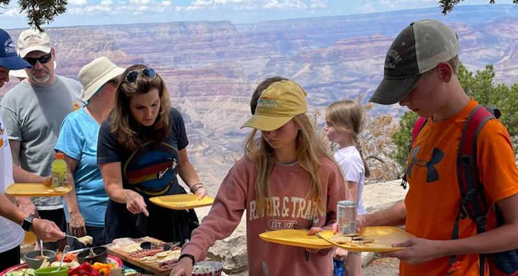 Les visiteurs servent des aliments de pique-nique sur des assiettes jaunes au bord d'un canyon, avec le Grand Canyon qui s'étend derrière eux.