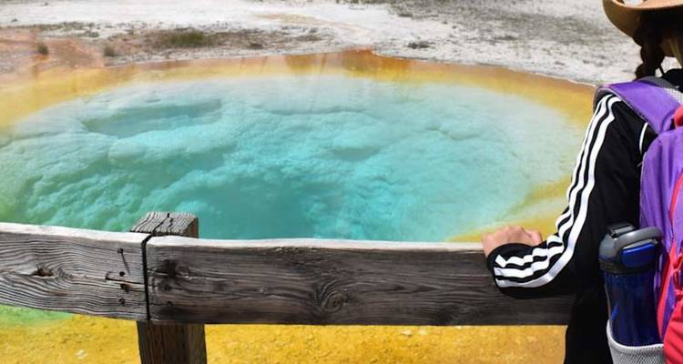 Un voyageur contemple une source thermale d'un bleu turquoise éclatant encadrée par des anneaux de minéraux orange et jaunes.