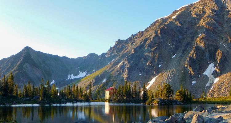 Un refuge alpin isolé se reflète dans un lac de montagne calme à l'heure dorée sous des pics rocheux escarpés.