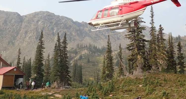 Un hélicoptère rouge survole un lodge de montagne isolé entouré de sapins et de montagnes.
