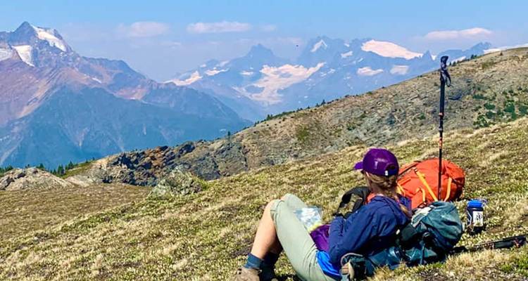 Un randonneur se repose sur une crête avec son sac à dos et son bâton de trekking, surplombant des sommets enneigés.