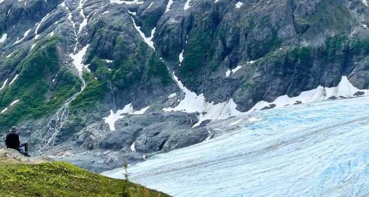 Le randonneur se repose sur une petite colline herbeuse à côté d'un vaste glacier bleu qui dévale les pics accidentés.