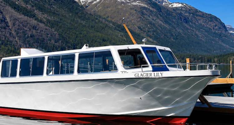 El barco turístico blanco 'Glacier Lily' está amarrado en un lago tranquilo con picos nevados de fondo.