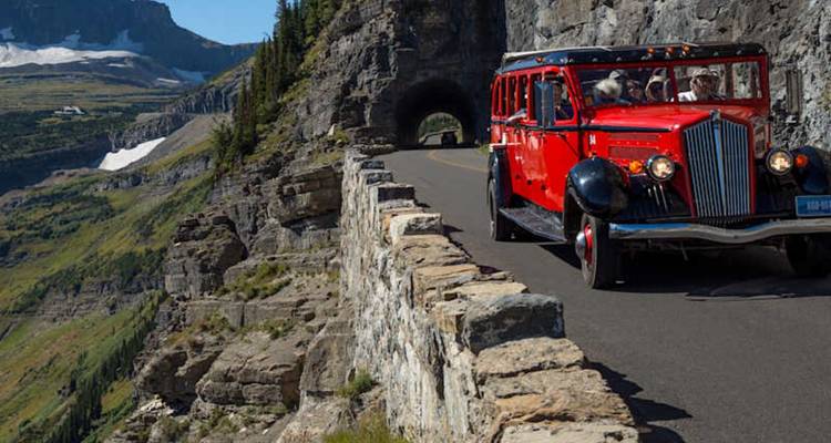 Un autobús turístico rojo histórico sale de un túnel de roca en una carretera de paso de montaña.