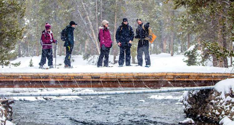 Des voyageurs vêtus de vêtements d'hiver se tiennent sur un pont enneigé surplombant un ruisseau qui coule.