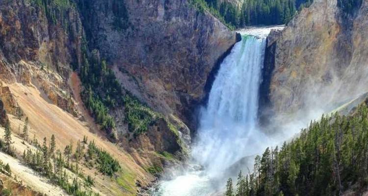 Une puissante cascade se précipite dans un canyon brumeux encadré par des falaises abruptes et des arbres à feuillage persistant.
