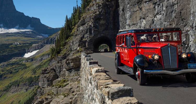 Un iconique bus vintage rouge circule le long d'une route surplombant les falaises, passant devant un tunnel dans une luxuriante vallée alpine.