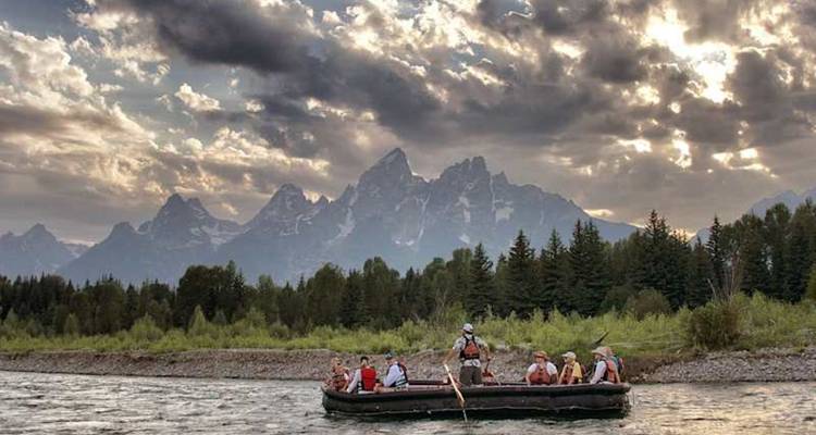 Un radeau transportant des touristes dérive sur la rivière avec les pics dentelés de la Teton se détachant en silhouette contre des nuages spectaculaires