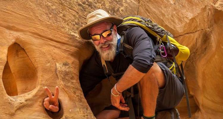 Un randonneur barbu fait un signe de paix en passant difficilement par un étroit trou de grès.