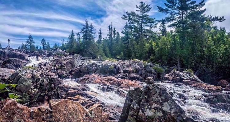 Cascade rocheuse sauvage avec des eaux tumultueuses entourée d'une forêt de pins sous un ciel bleu zébré